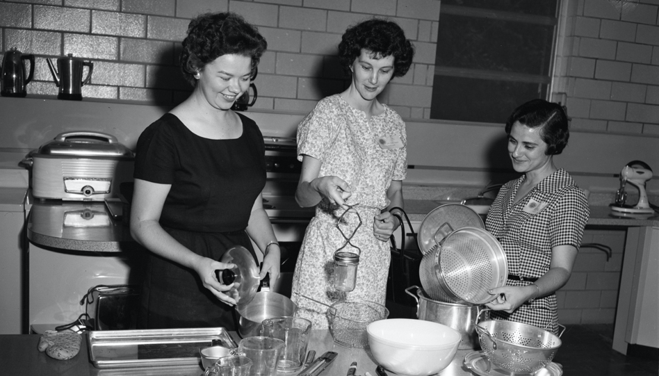 Black-and-white photo of women standing in a kitchen organizing pots and pans